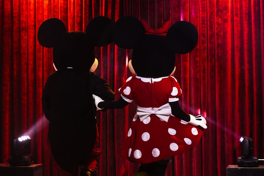 Mickey e Minnie em palco, de costas, frente a uma cortina vermelha